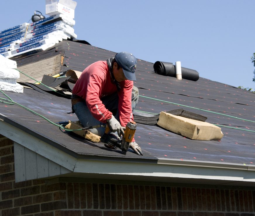 A roofer fastening shingles along the eaves of a house.